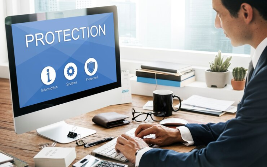 A professional in a dark suit typing on a computer with a screen displaying the word 'PROTECTION' alongside icons representing information, systems, and security.