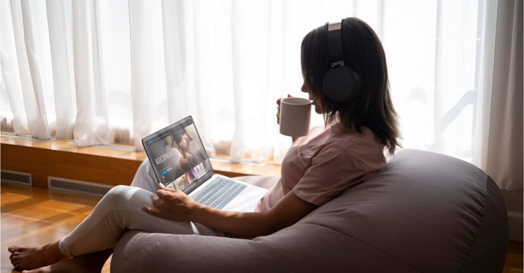 Woman sitting comfortably on a bean bag, wearing headphones, and watching a video on a laptop, symbolizing the ease of access provided by private video hosting platforms.
