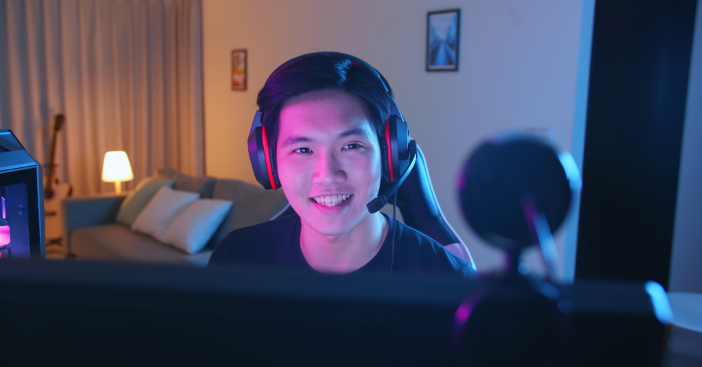 A young man with headphones live streaming a gaming session in a well-lit room at night.