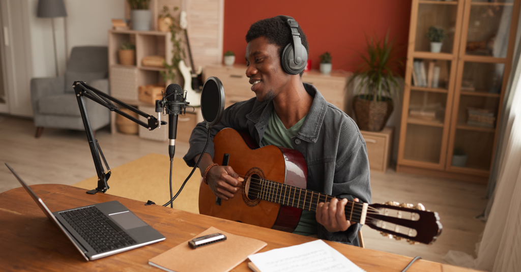A musician live streaming an acoustic guitar session from home with a microphone and laptop setup.